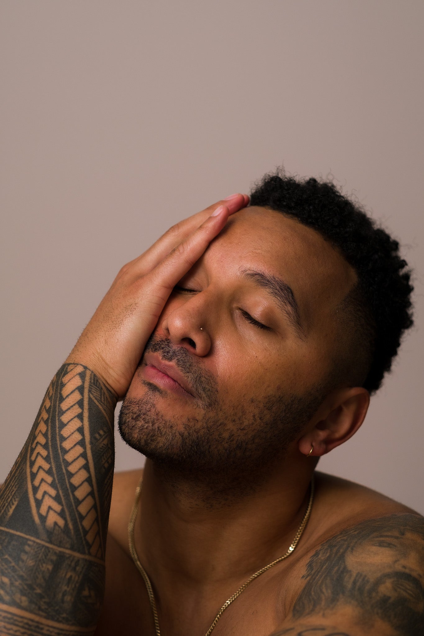 A close-up of a tattooed man, wearing Seven:Thirty products, shown from the shoulders up. His hand is resting on his face, with his eyes closed, against a khaki background.