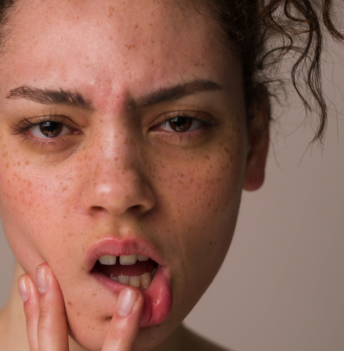 A close-up of a woman's face, wearing Seven:Thirty products, against a khaki background. Her mouth is slightly open, with her finger pressed against her bottom lip.