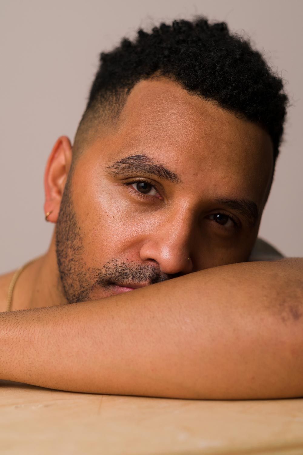 A close-up of a man wearing Seven:Thirty products, resting his face on his forearm, against a khaki background.