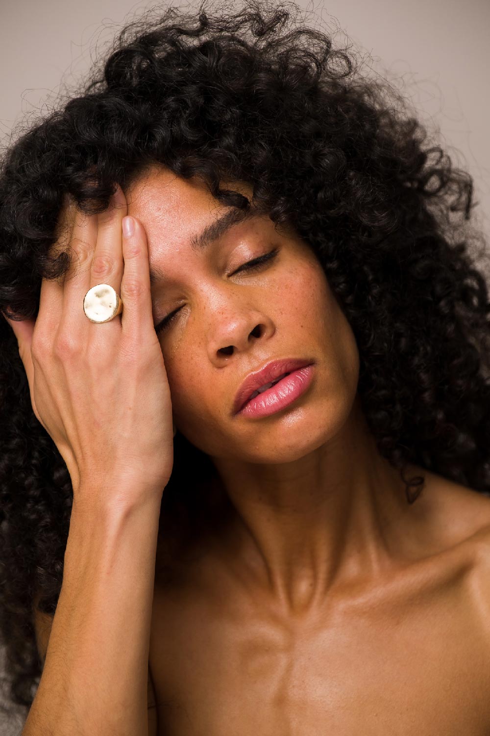 A close-up of a woman's face, from the décolletage up, wearing Seven:Thirty products, against a khaki background. Her eyes are closed and one hand is touching her forehead.