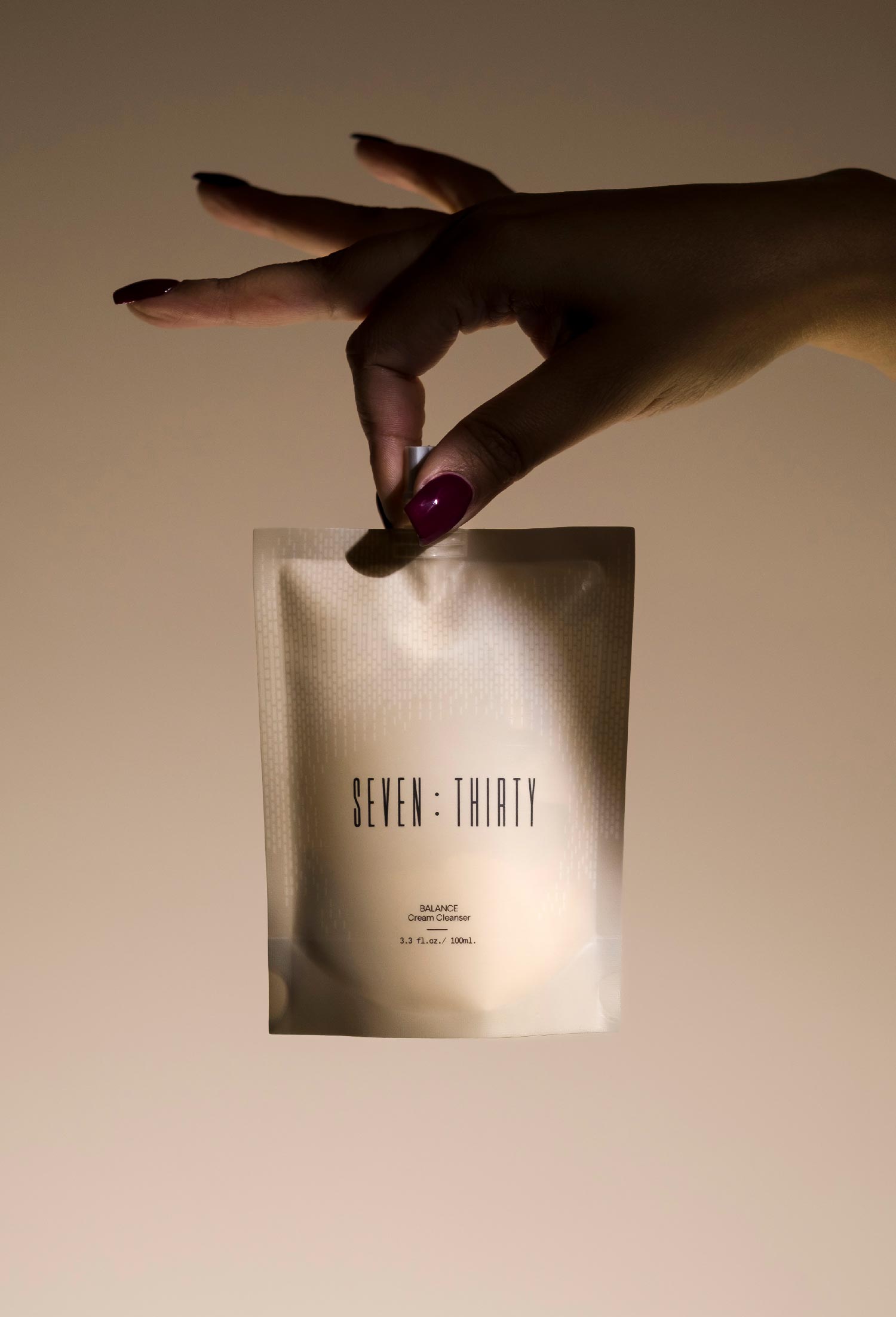 A hand with dark red nails holding up the BALANCE Cream Cleanser by her thumb and index finger against a khaki background