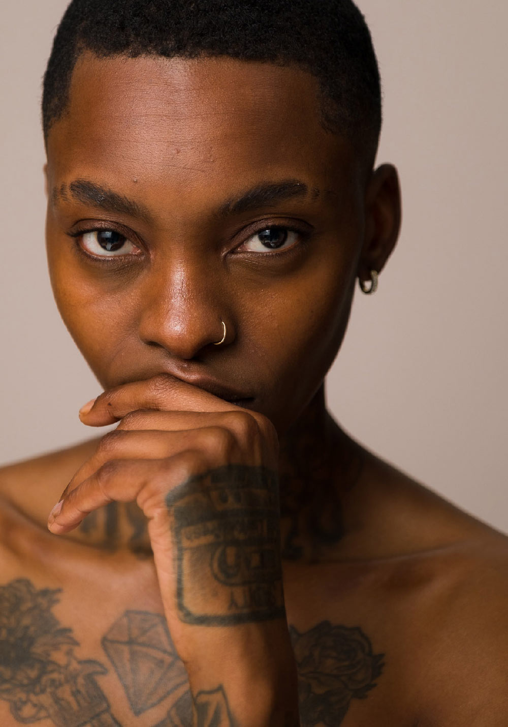 A close-up of a woman's face, from the décolletage up, wearing Seven:Thirty products, against a khaki background. She has tattoos on her chest and hand, with that hand covering her lips.