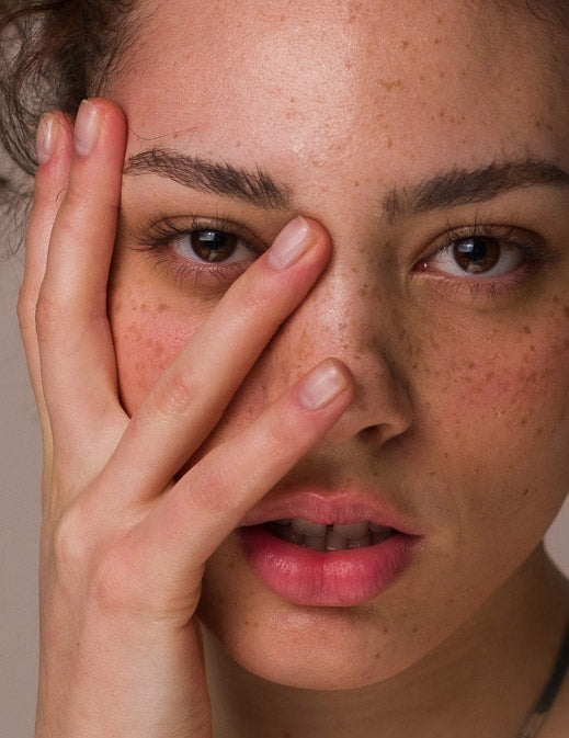 A close-up of a woman's face, wearing Seven:Thirty products, with the fingers of one hand touching her face. She has freckles and her lips are slightly parted.