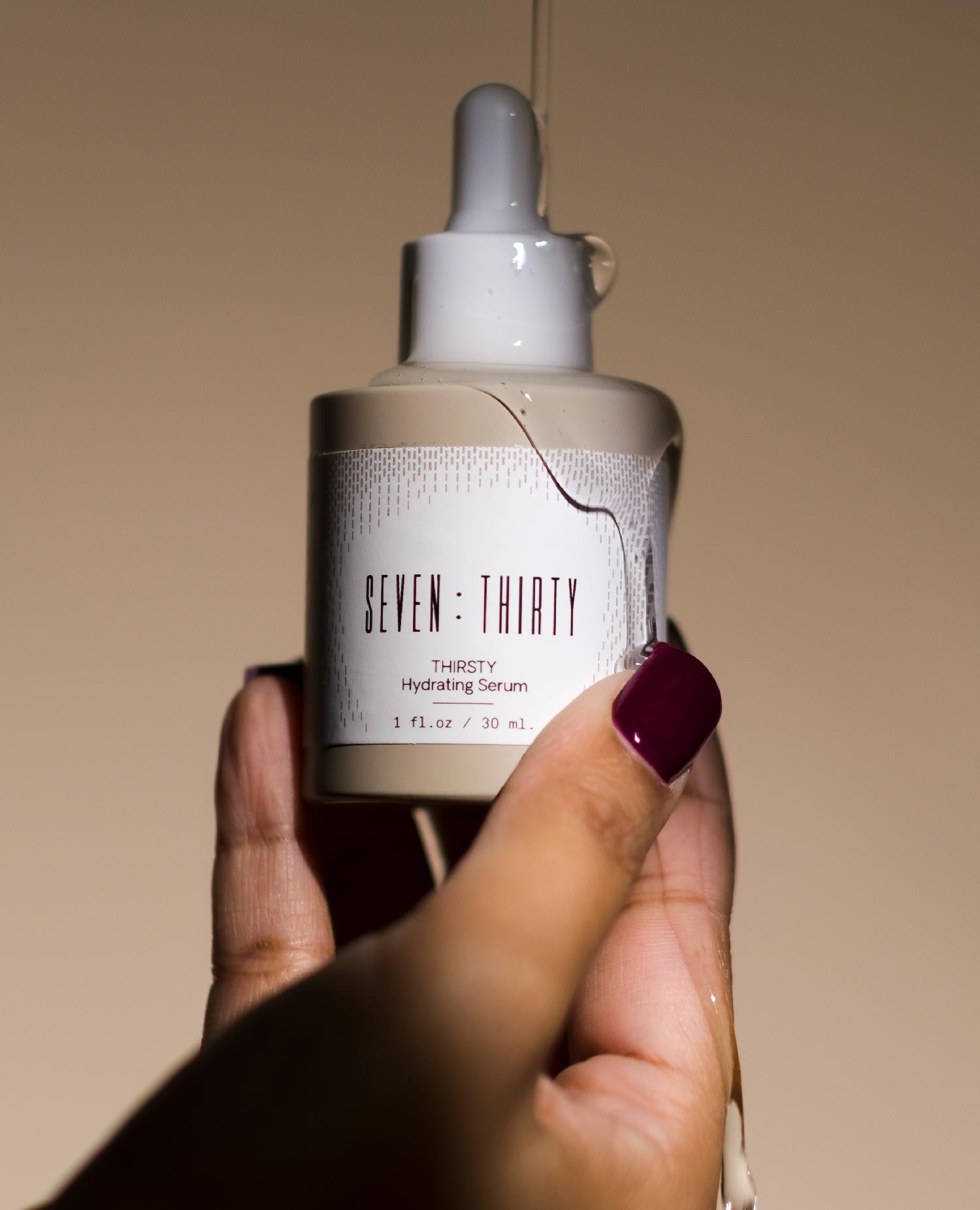 A hand with dark red nails holding THIRSTY Hydrating Serum against a khaki background with liquid product dripping onto the bottle and fingers.