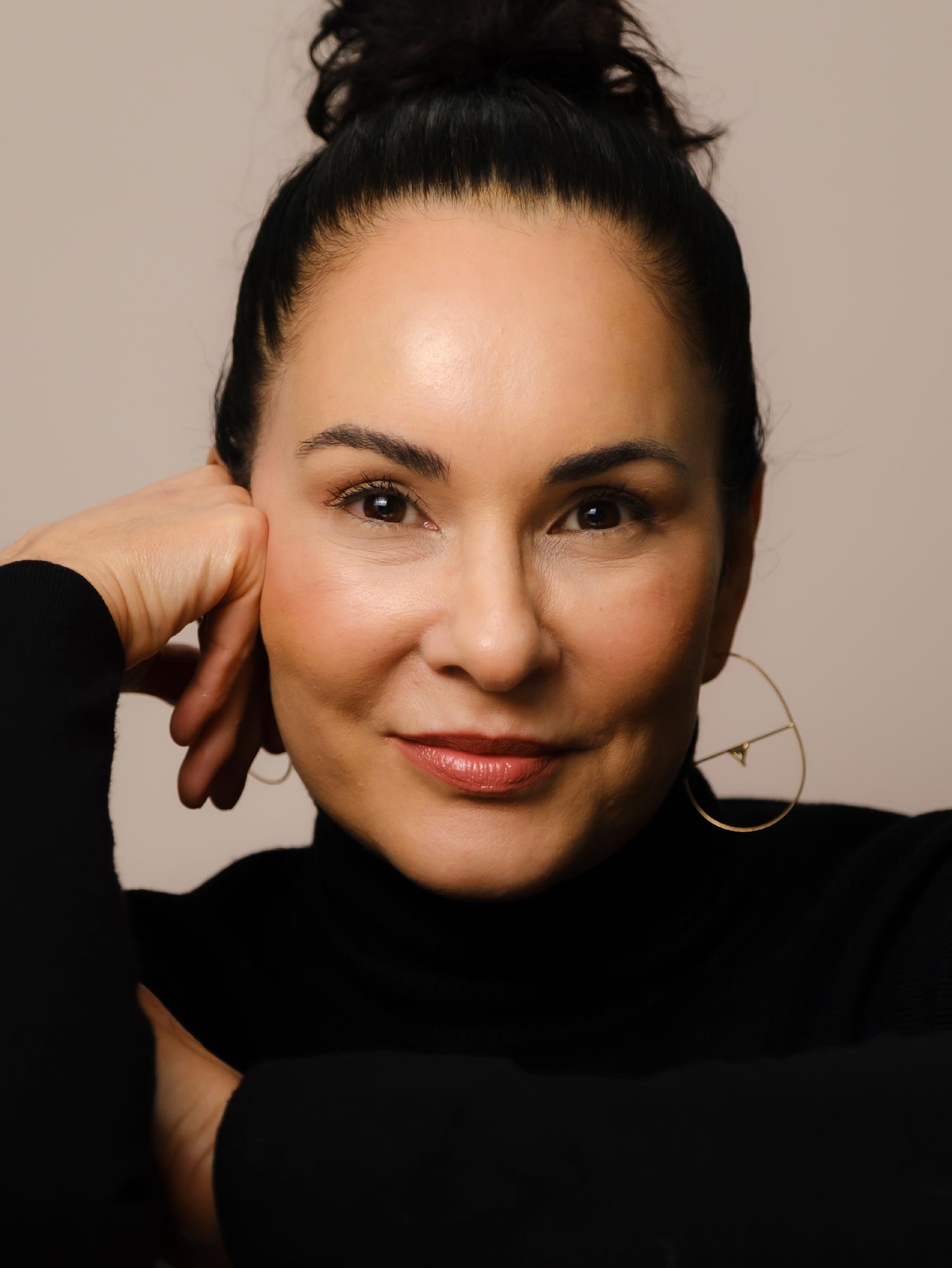 A close-up portrait of Shannon Carter-Gascon, wearing a black turtleneck with gold hoop earrings, against a khaki background.